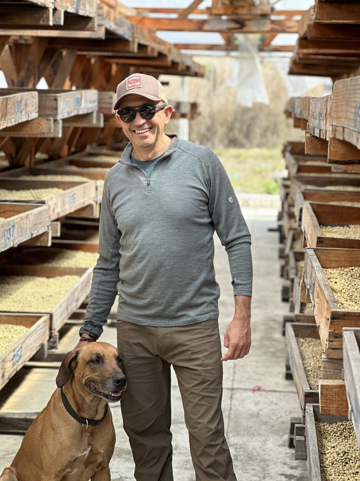 A smiling man in a cap with his dog among wooden raised drying racks of parchment coffee at Finca Santa Ana