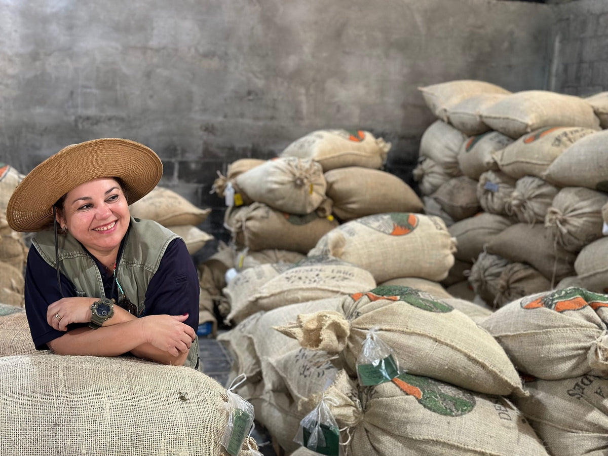 A smiling woman in a sun hat leaning on stacked burlap coffee sacks in the Finca Las Mercedes warehouse