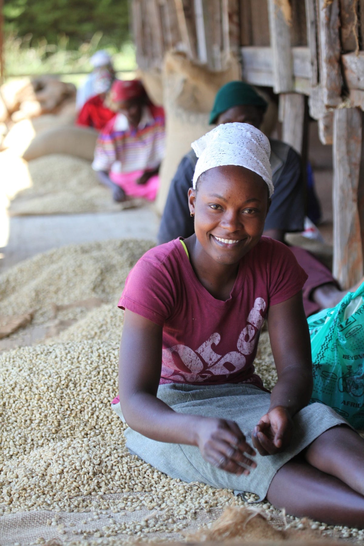 Young woman in a white headscarf smiling at the camera while hand-sorting green coffee in a warehouse