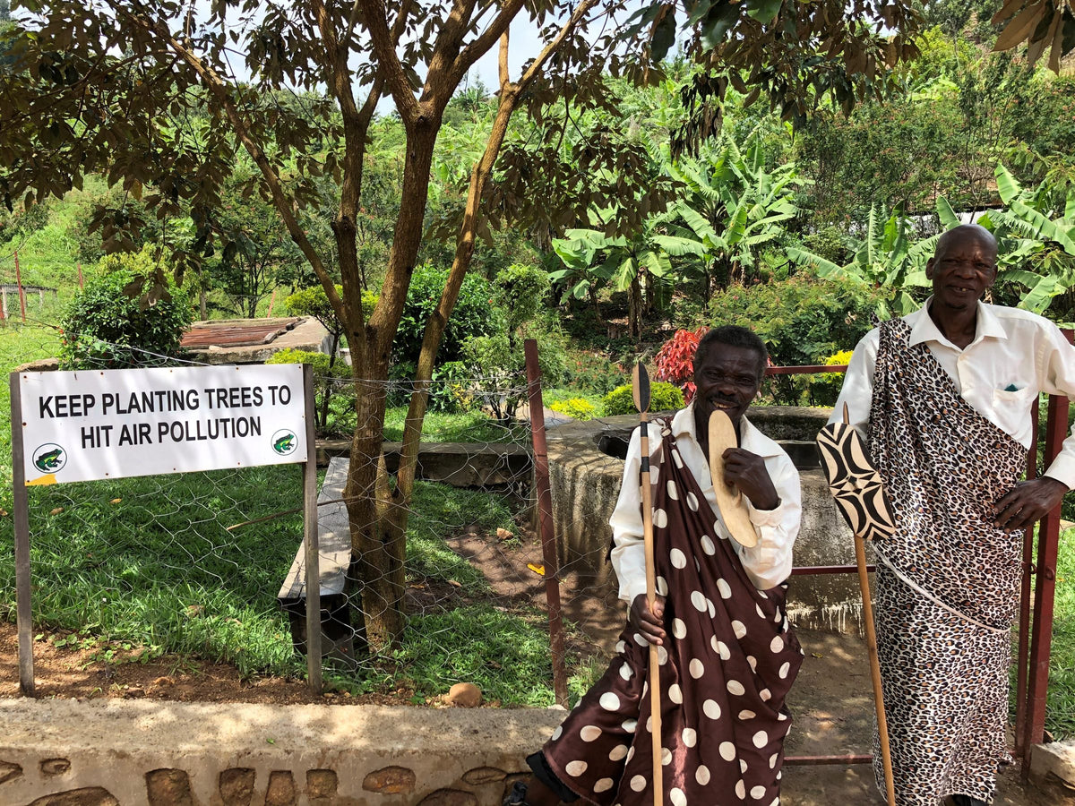 Two elders in traditional patterned wraps standing under trees beside a 'Keep planting trees' sign